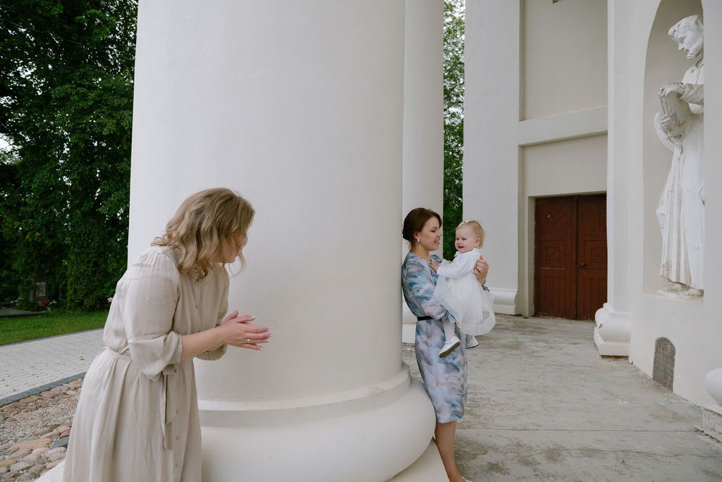 Two women and a baby pose near white columns of an elegant building surrounded by lush greenery.