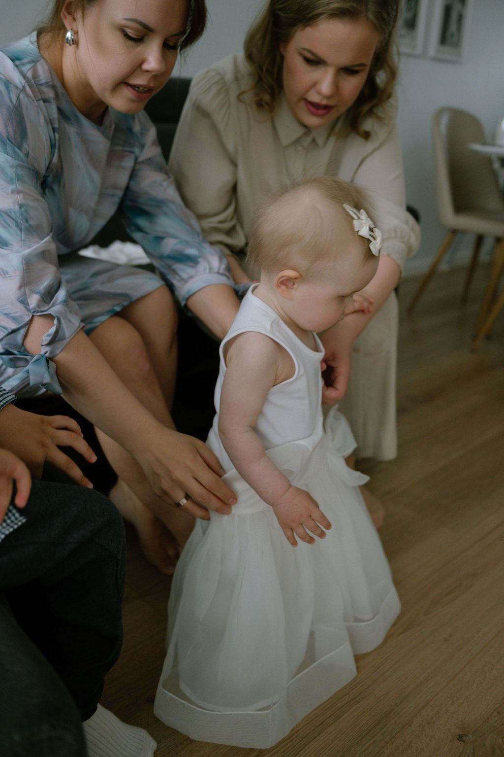 Two women helping a baby girl in a white dress take her first steps on a wooden floor.