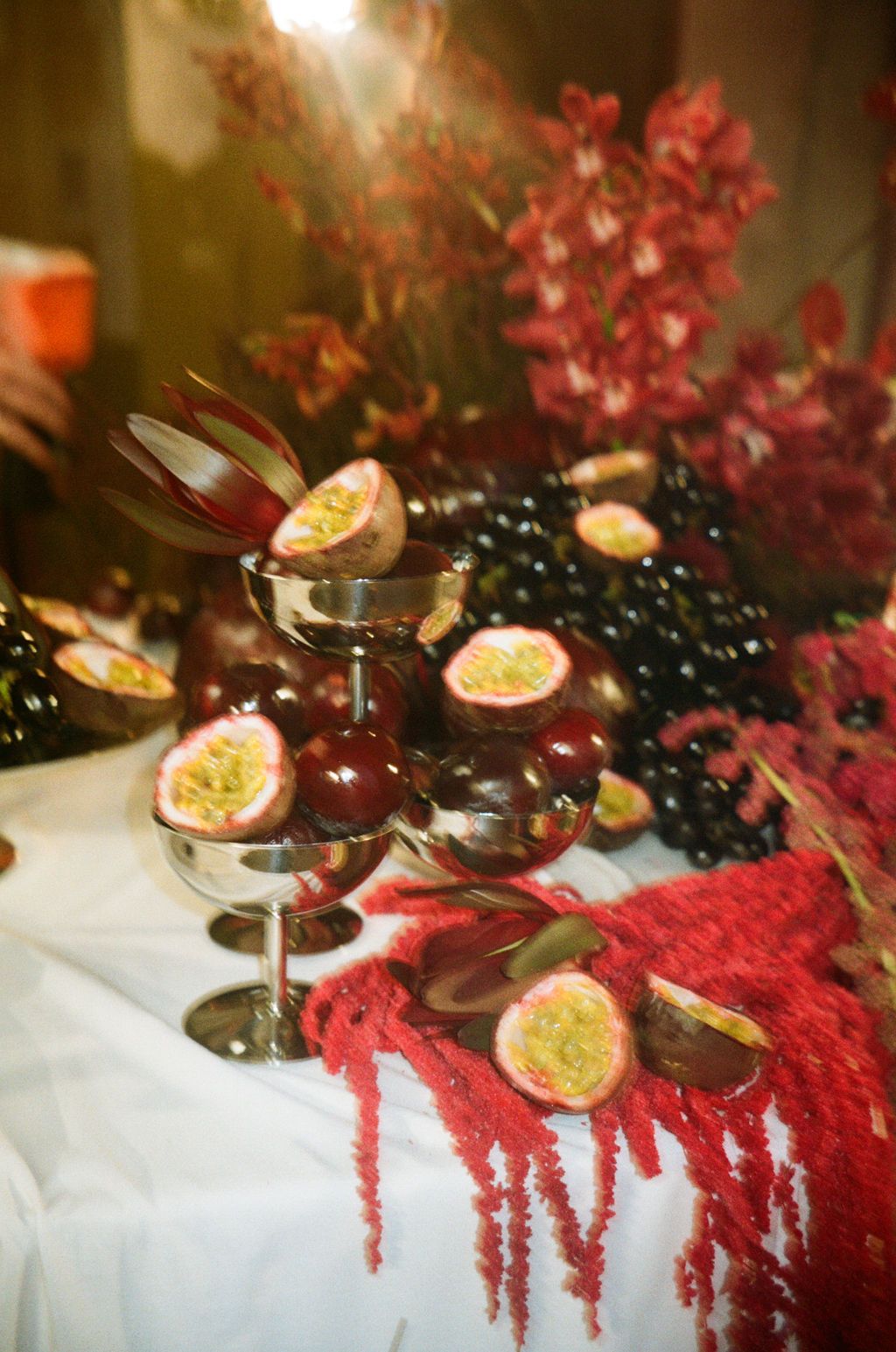 Elegant fruit display with passion fruit, dark grapes, and red flowers arranged on silver stands and a white tablecloth.