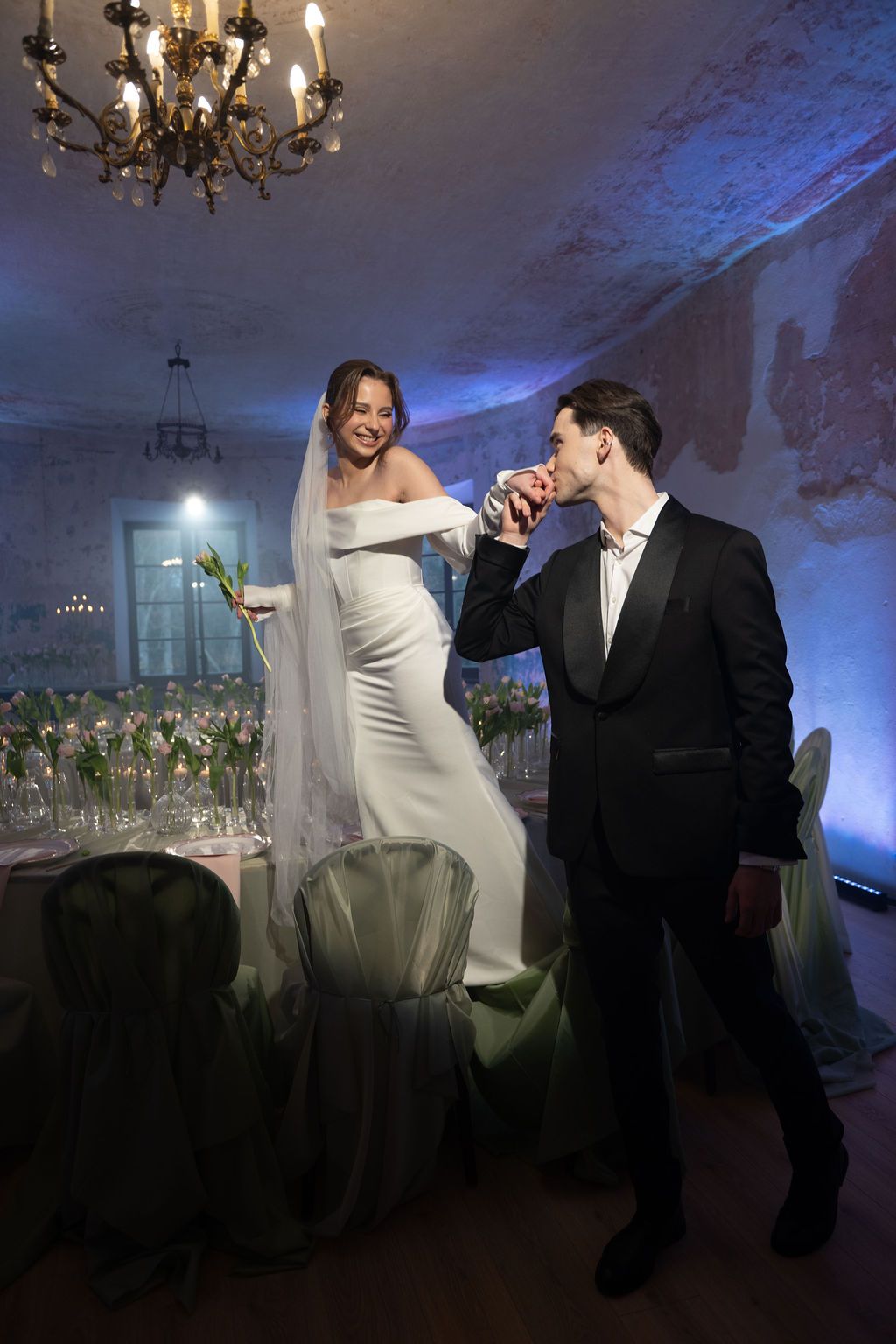 Groom in black tuxedo kissing bride's hand at elegant wedding reception with chandelier and blue lighting.