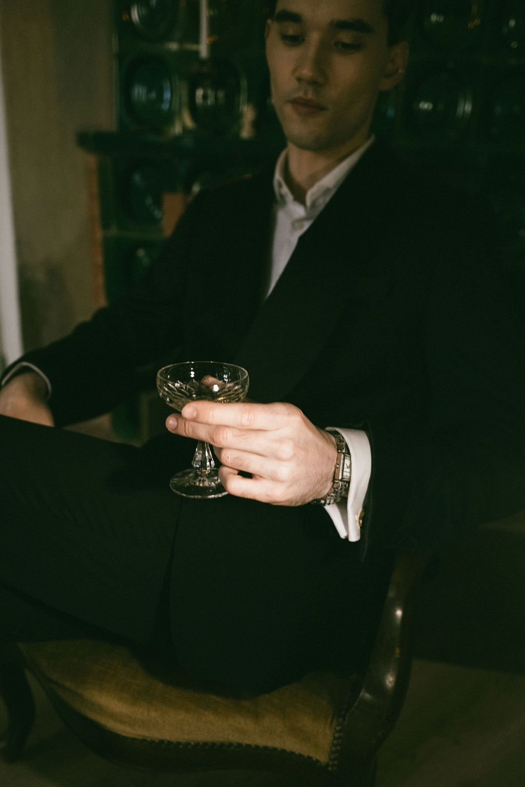 Elegant man in black suit holding a crystal cocktail glass while seated in a vintage chair.