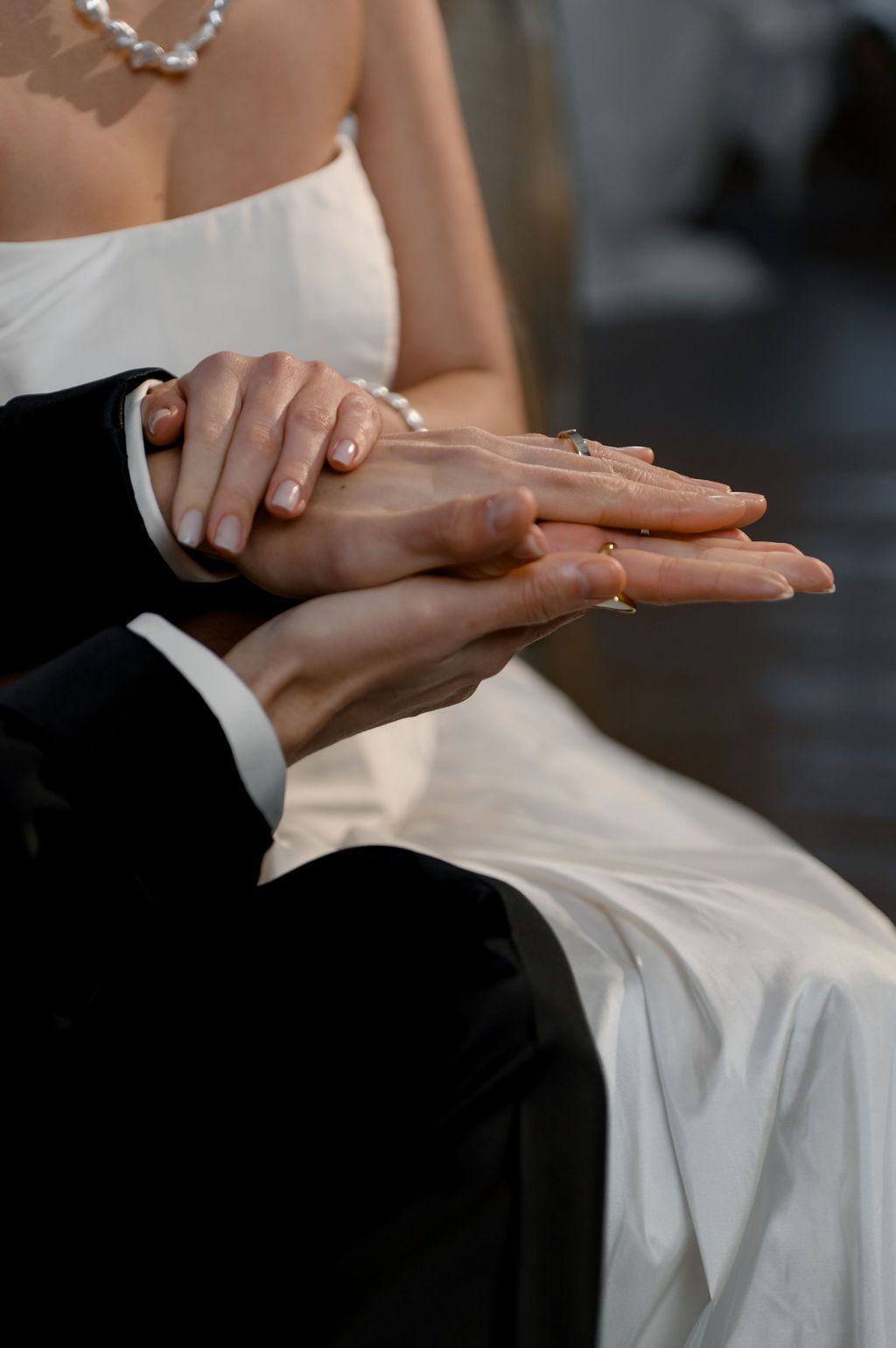 Bride and groom holding hands at wedding ceremony, showing pearl bracelet and wedding rings in close-up shot.
