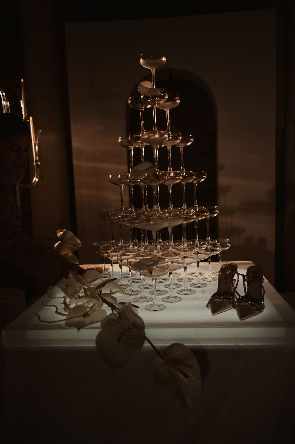Elegant champagne tower with stacked glasses on illuminated table in dimly lit room with heels and flowers.
