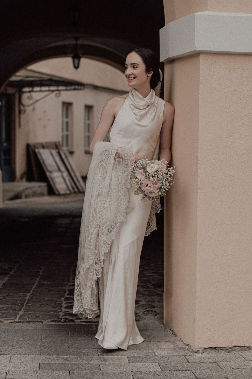 Bride in ivory satin halter gown with lace shawl holding pink bouquet, leaning against a peach column.
