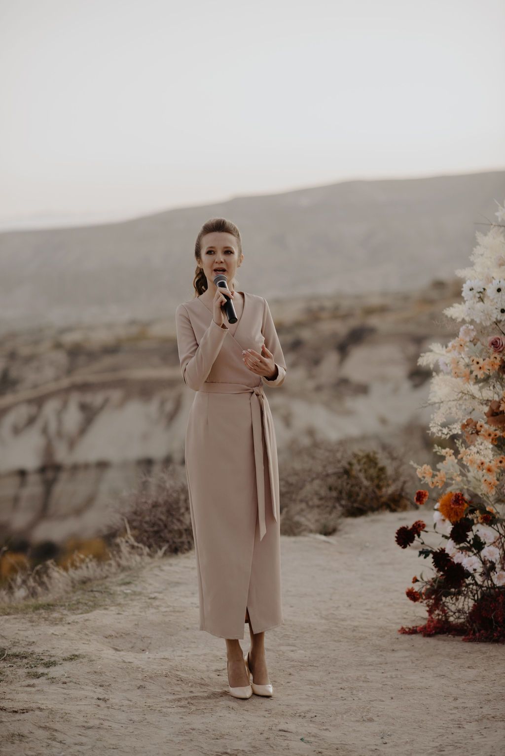 Woman in beige midi dress holding microphone outdoors in desert landscape with floral arrangement nearby.