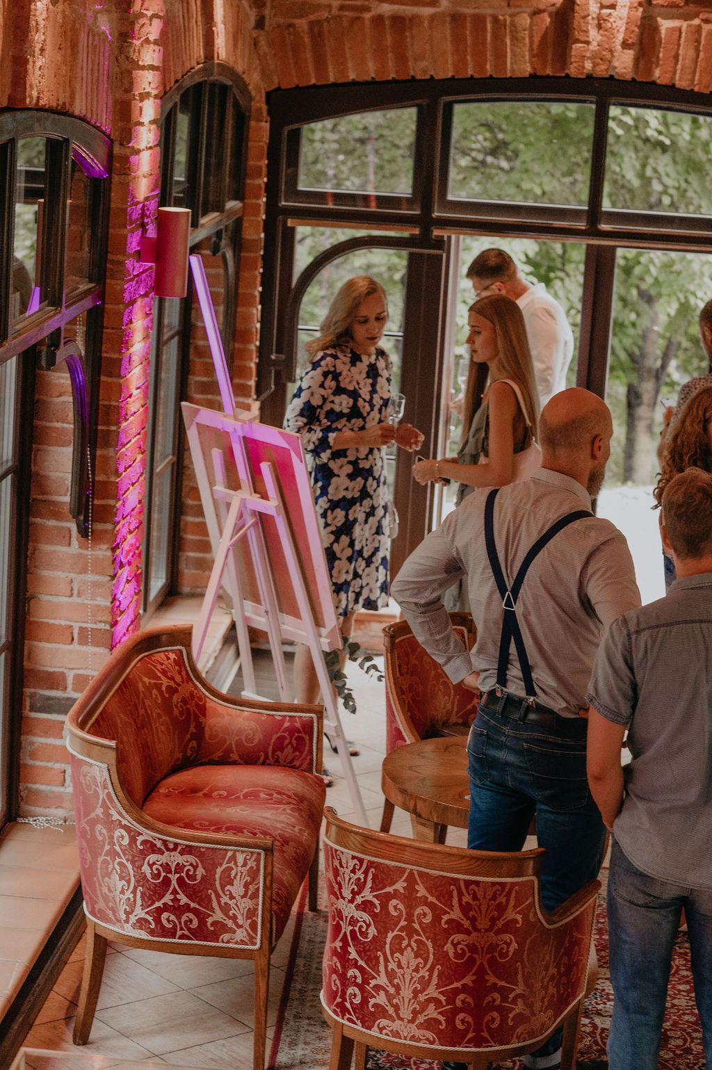 Guests gather around a pink neon art easel in an elegant brick venue with ornate red velvet chairs.