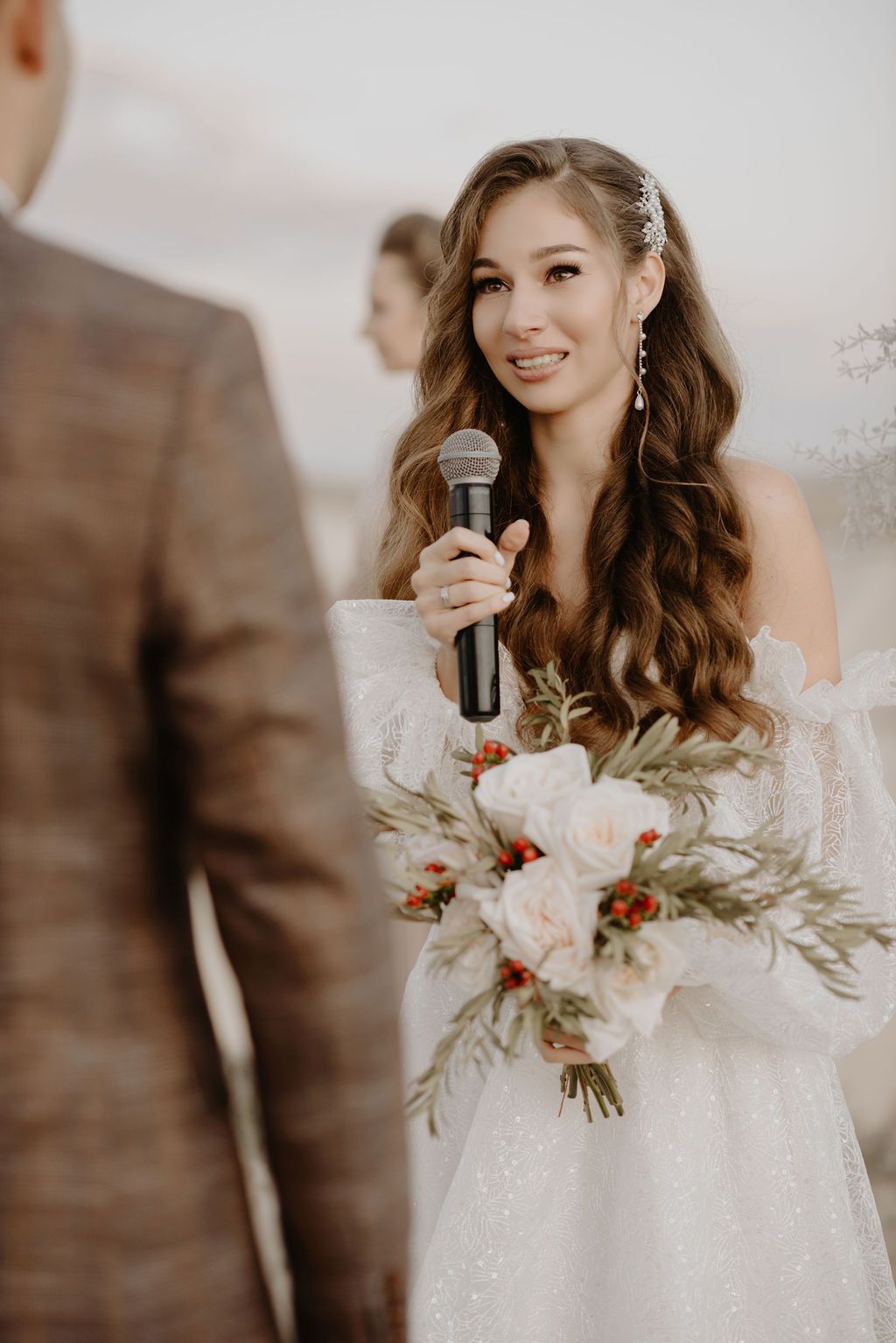 Smiling bride in white off-shoulder gown holding microphone and floral bouquet during wedding ceremony.