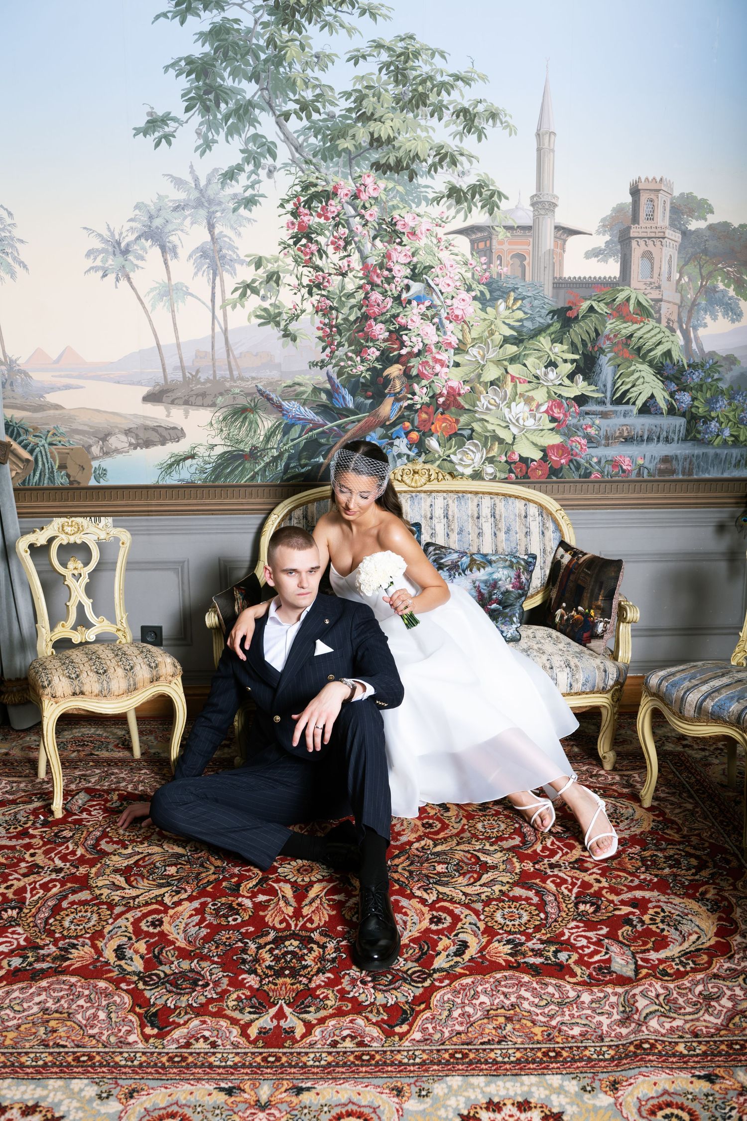Elegant bride and groom posing in ornate room with floral mural, antique furniture, and red Persian rug.