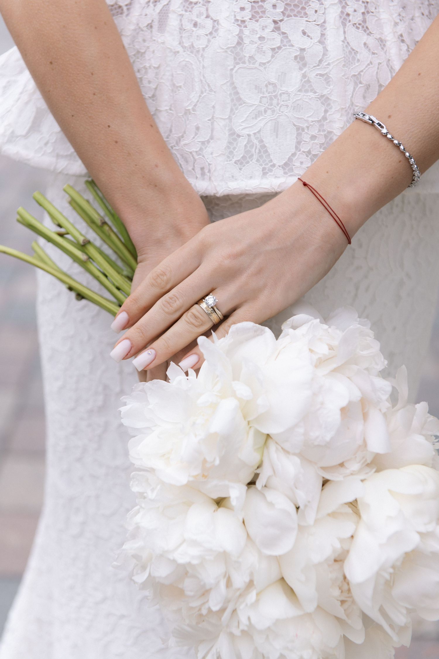 Bride holding white peony bouquet, wearing wedding ring and bracelet, in lace dress.