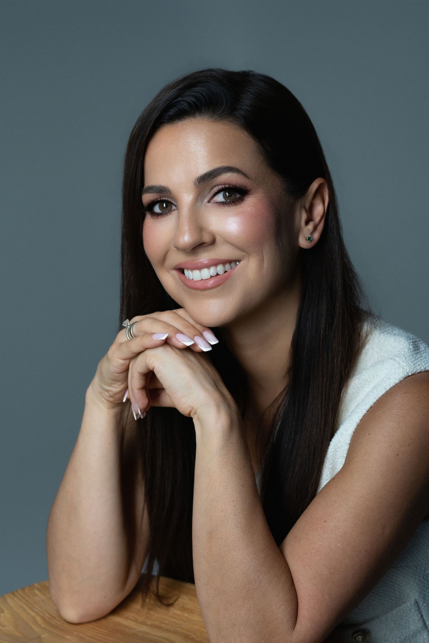 Smiling brunette woman with long hair poses with hands clasped, wearing white top against gray background.