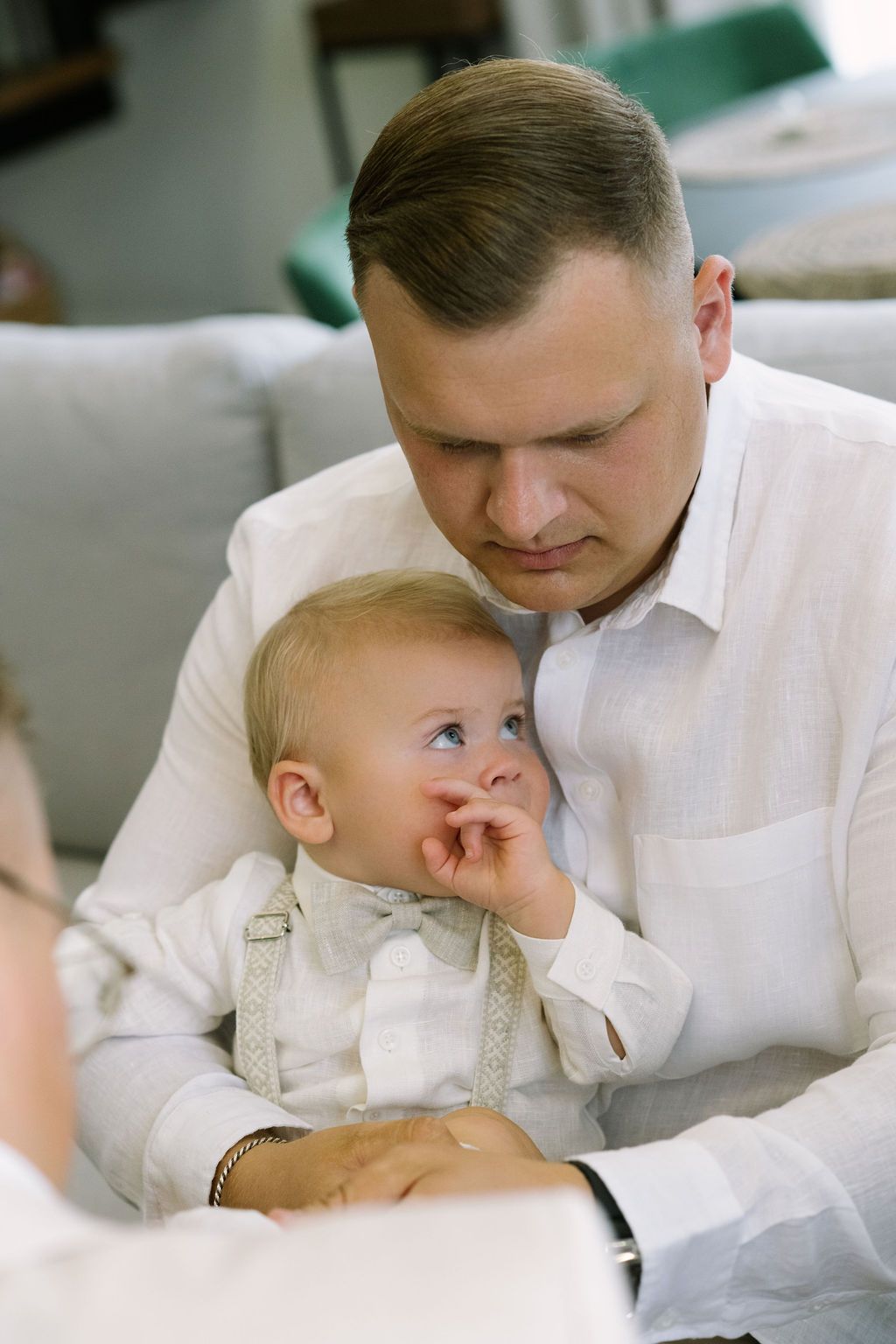 Loving father in white shirt cuddles smiling baby in white suspender outfit on a light sofa.