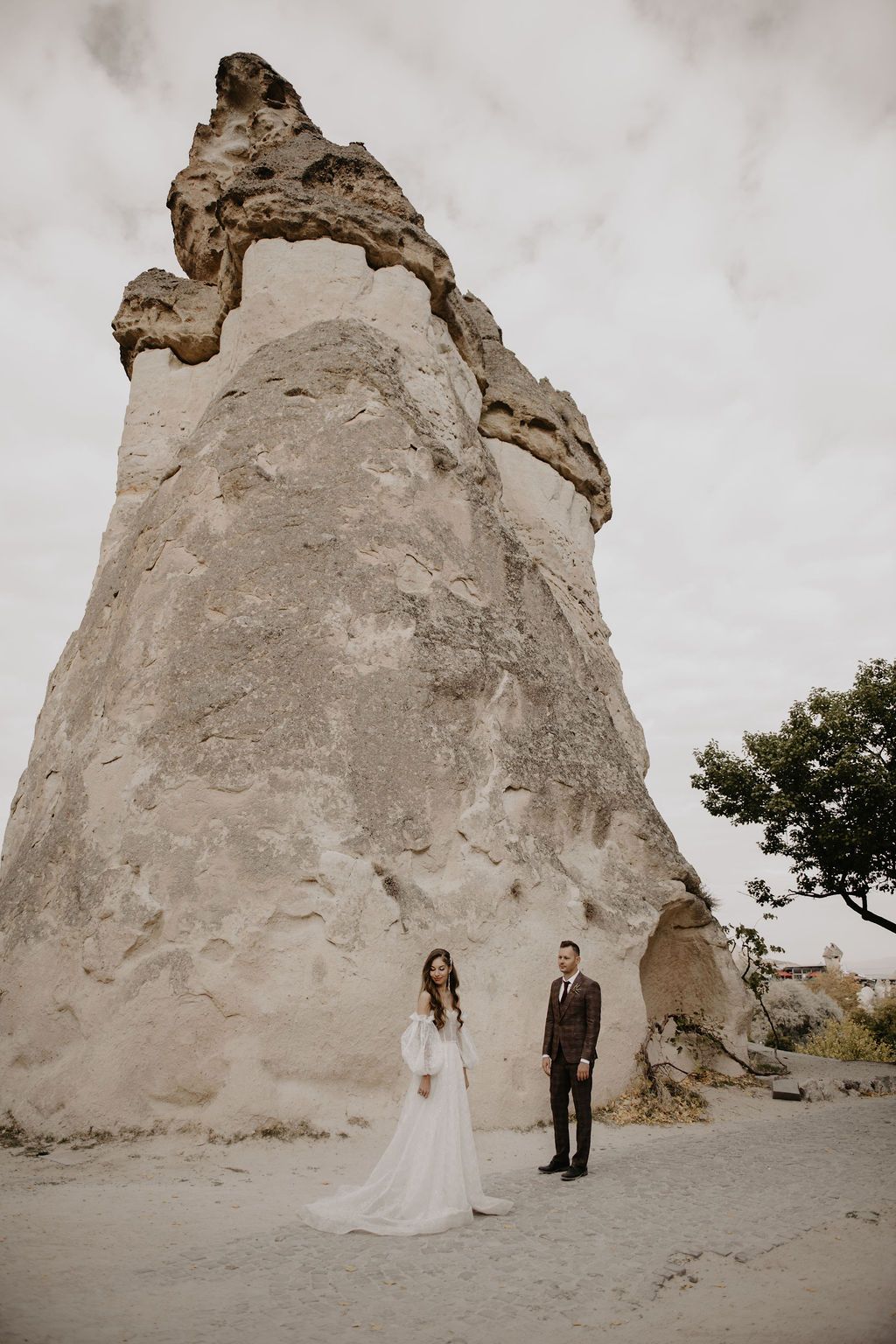 Bride and groom standing before a towering fairy chimney rock formation in Cappadocia, Turkey.