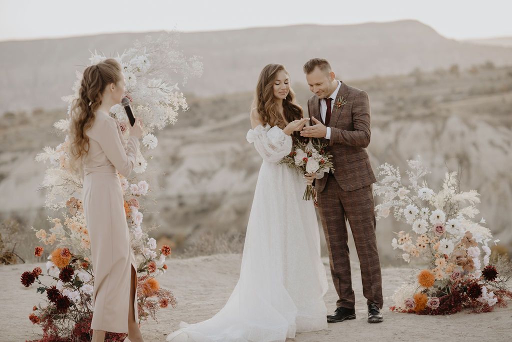 Outdoor desert wedding ceremony with bride in white gown, groom in plaid suit, and officiant beside floral arch.