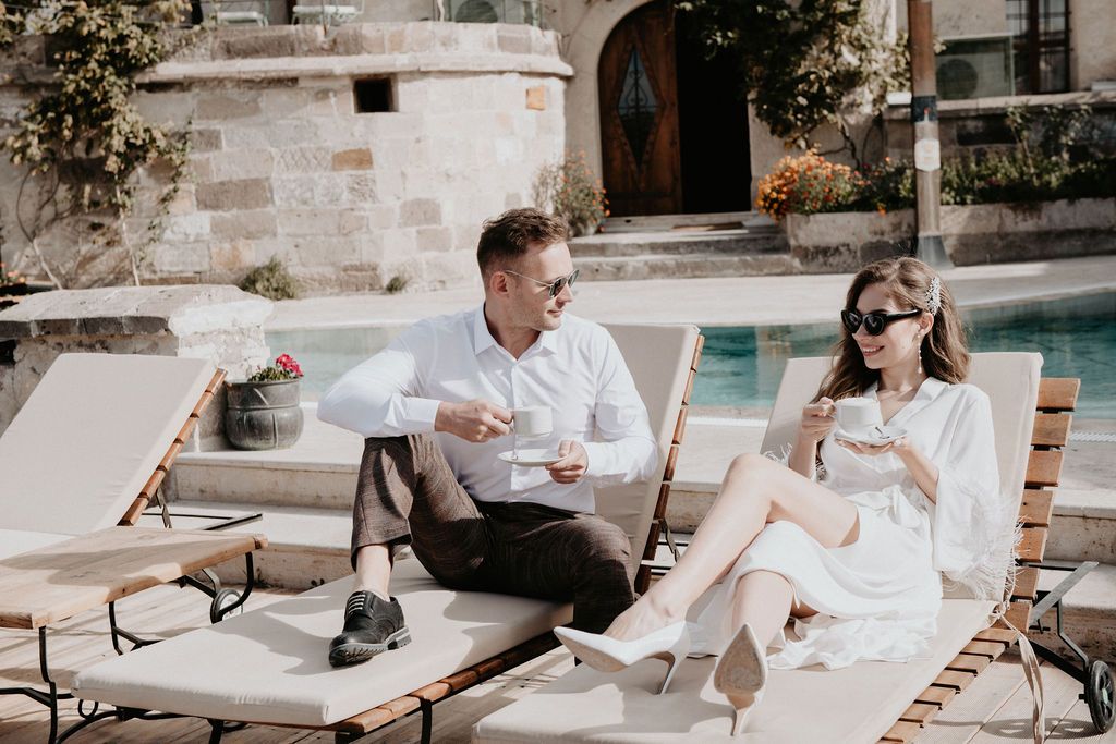 Couple relaxing on lounge chairs by a pool, enjoying drinks in a sunny outdoor setting near a stone building.