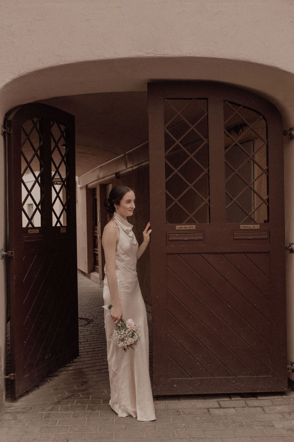 Bride in elegant ivory gown holding bouquet stands in arched wooden doorway of historic European building.