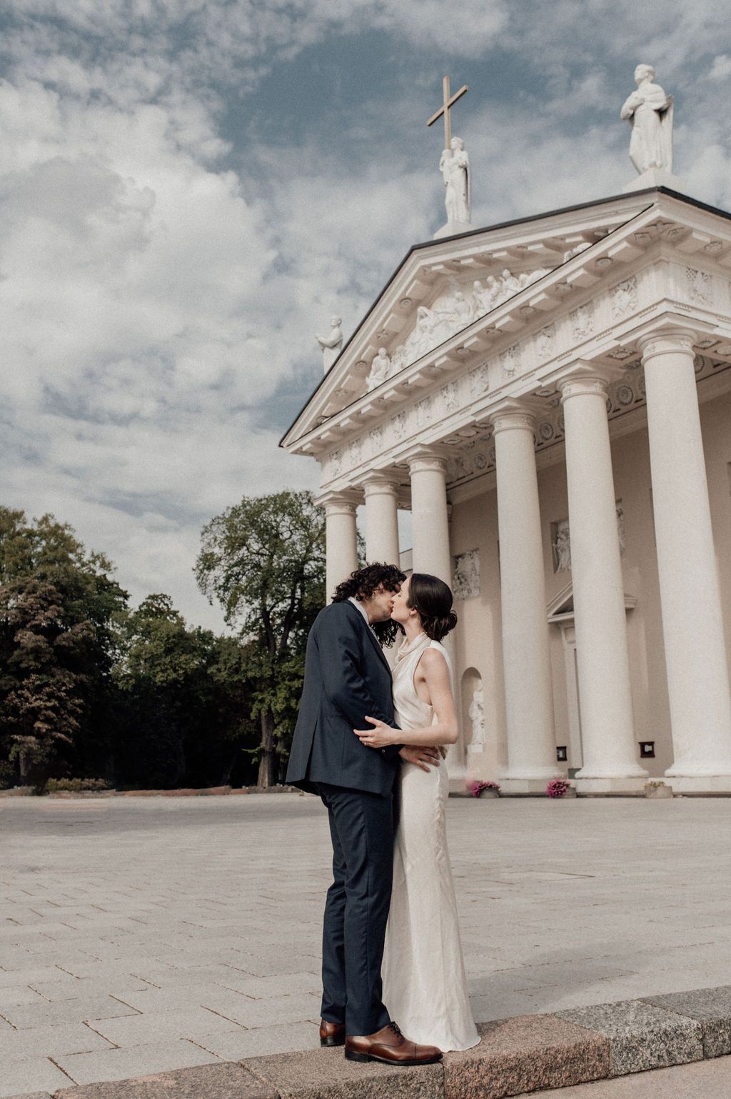 A couple shares a kiss in wedding attire before a grand white neoclassical cathedral with columns and statues.