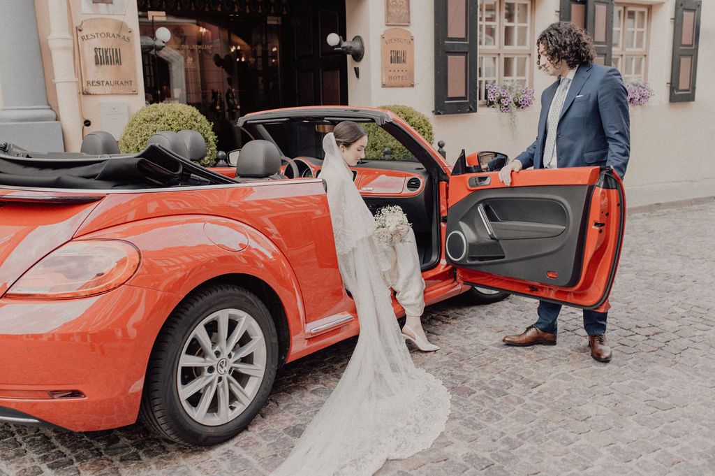 Bride in white gown exits red VW Beetle convertible as groom holds door open on cobblestone street.