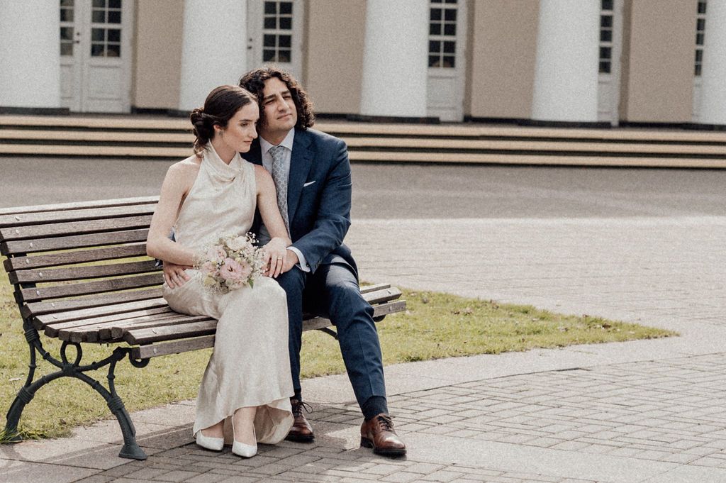 A bride and groom sit closely together on a park bench, dressed in wedding attire, sharing a romantic moment outdoors.