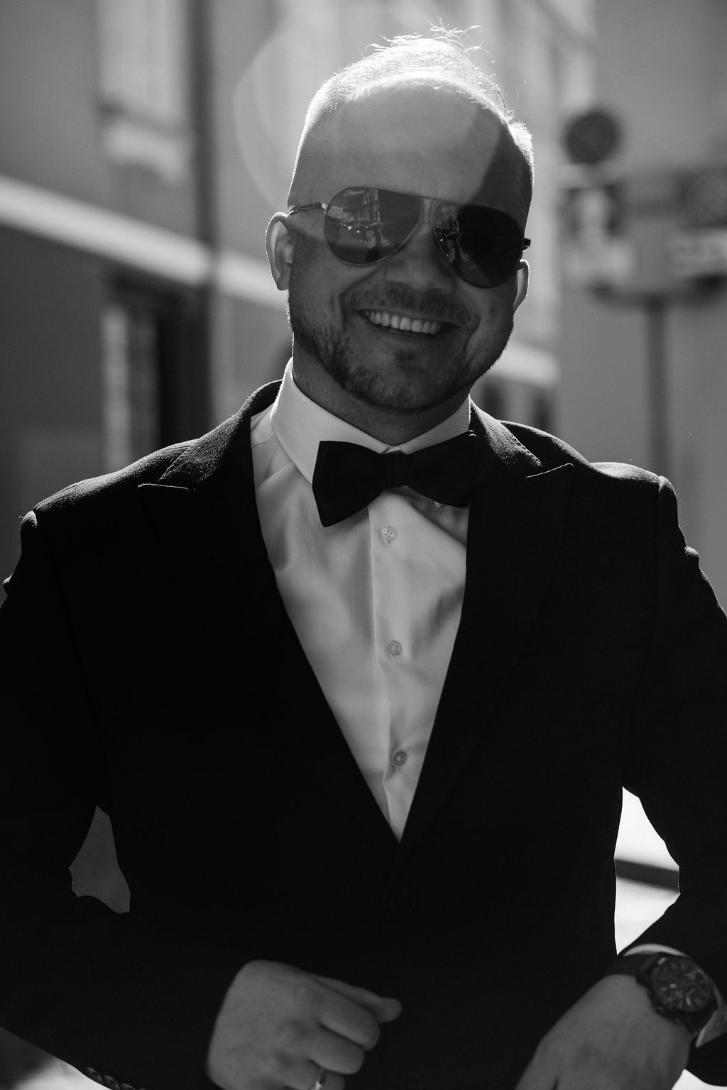 Smiling bald man in tuxedo with bow tie and aviator sunglasses poses outdoors in black and white photo.