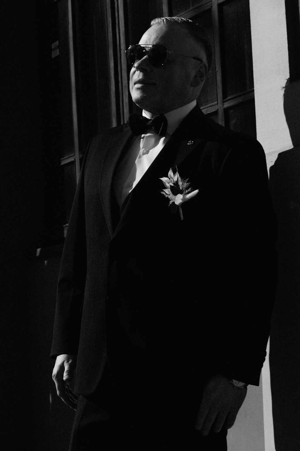 Elegant man in tuxedo with bow tie, boutonniere, and sunglasses poses in dramatic black and white portrait.