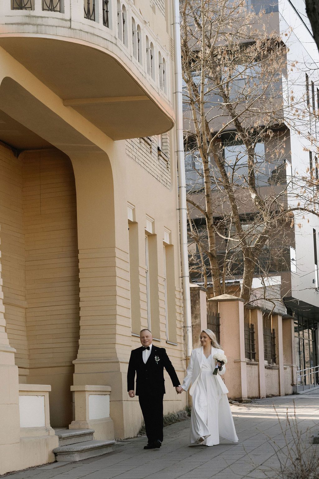Bride and groom walking hand in hand past an elegant cream building on their wedding day.