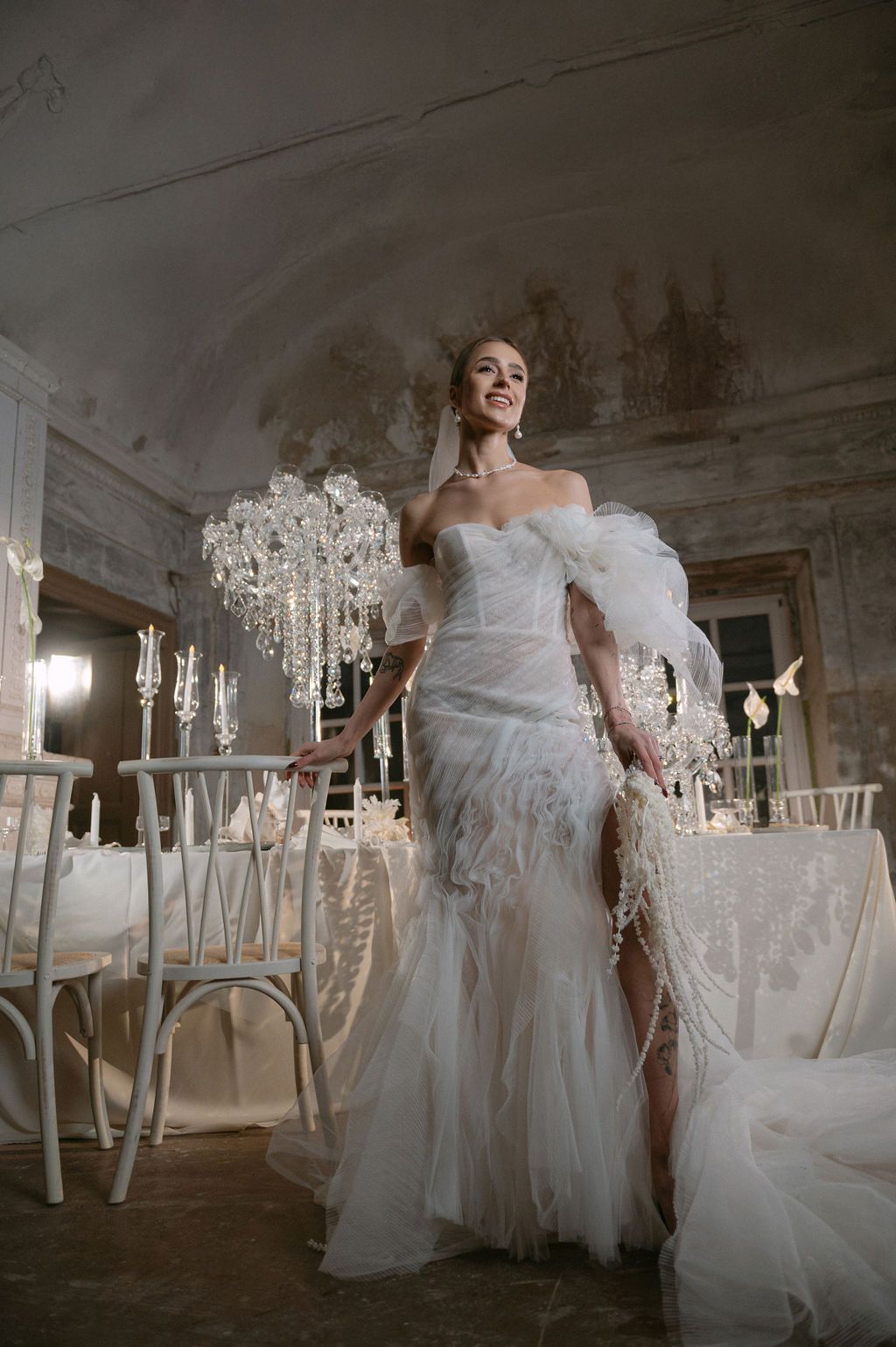 Elegant bride in a white feathered tulle gown poses in a luxurious crystal chandelier-adorned wedding venue.