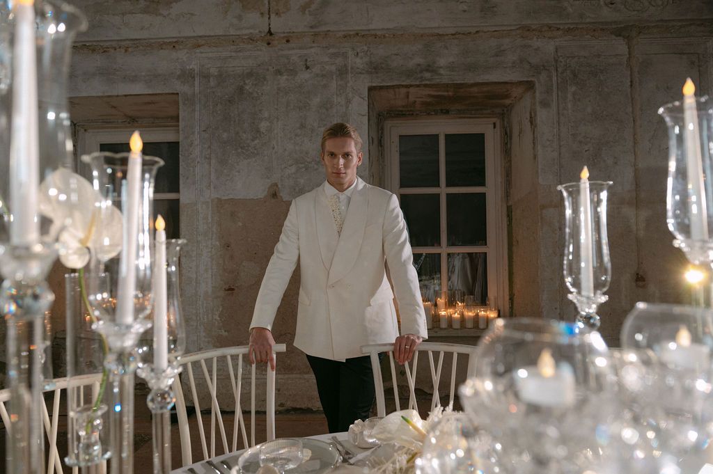 Elegant man in white jacket stands among crystal candelabras at a beautifully set dining table in a rustic venue.