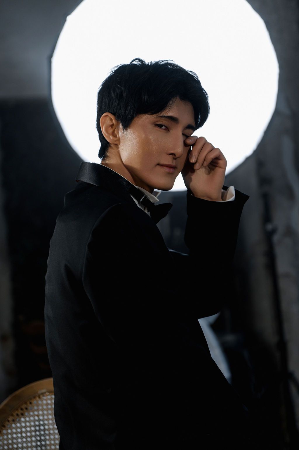 A young Asian man in a black suit poses thoughtfully against a bright circular light backdrop.