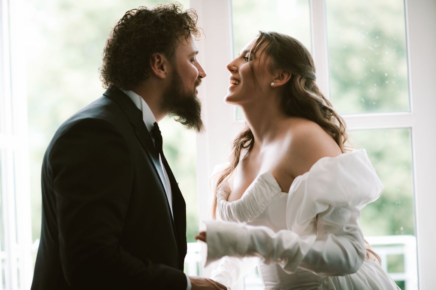 A bearded groom in a tuxedo and bride in an off-shoulder white dress share a joyful moment by a bright window.