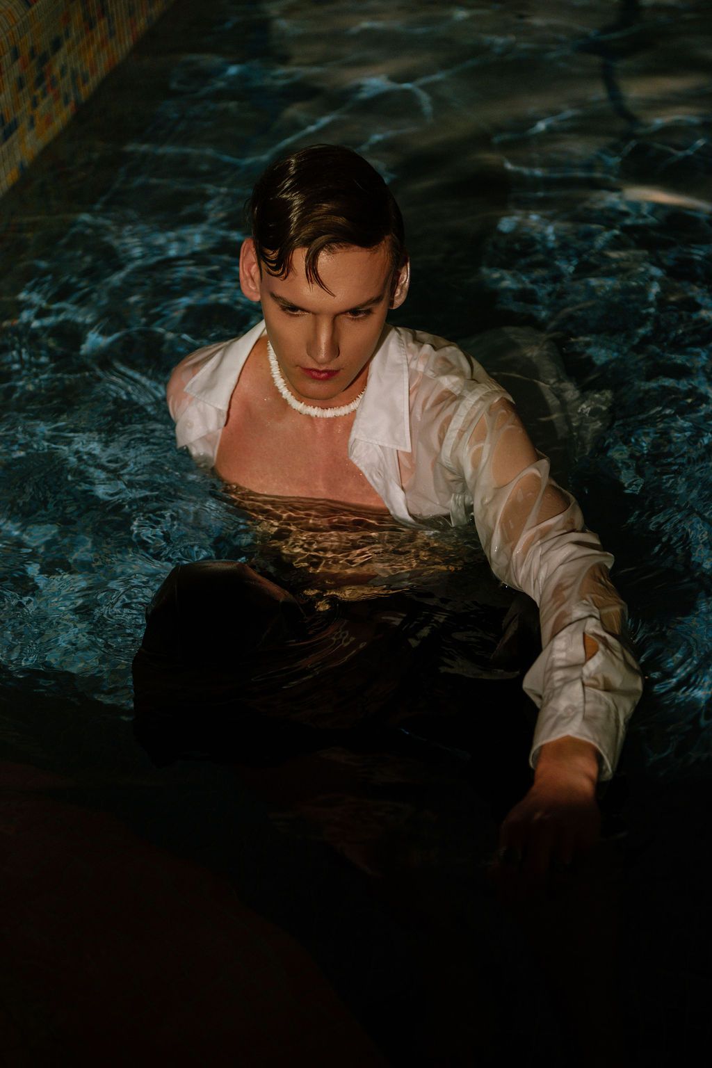 Young man in a wet white shirt and pearl necklace sits waist-deep in a dimly lit pool with rippling blue water.