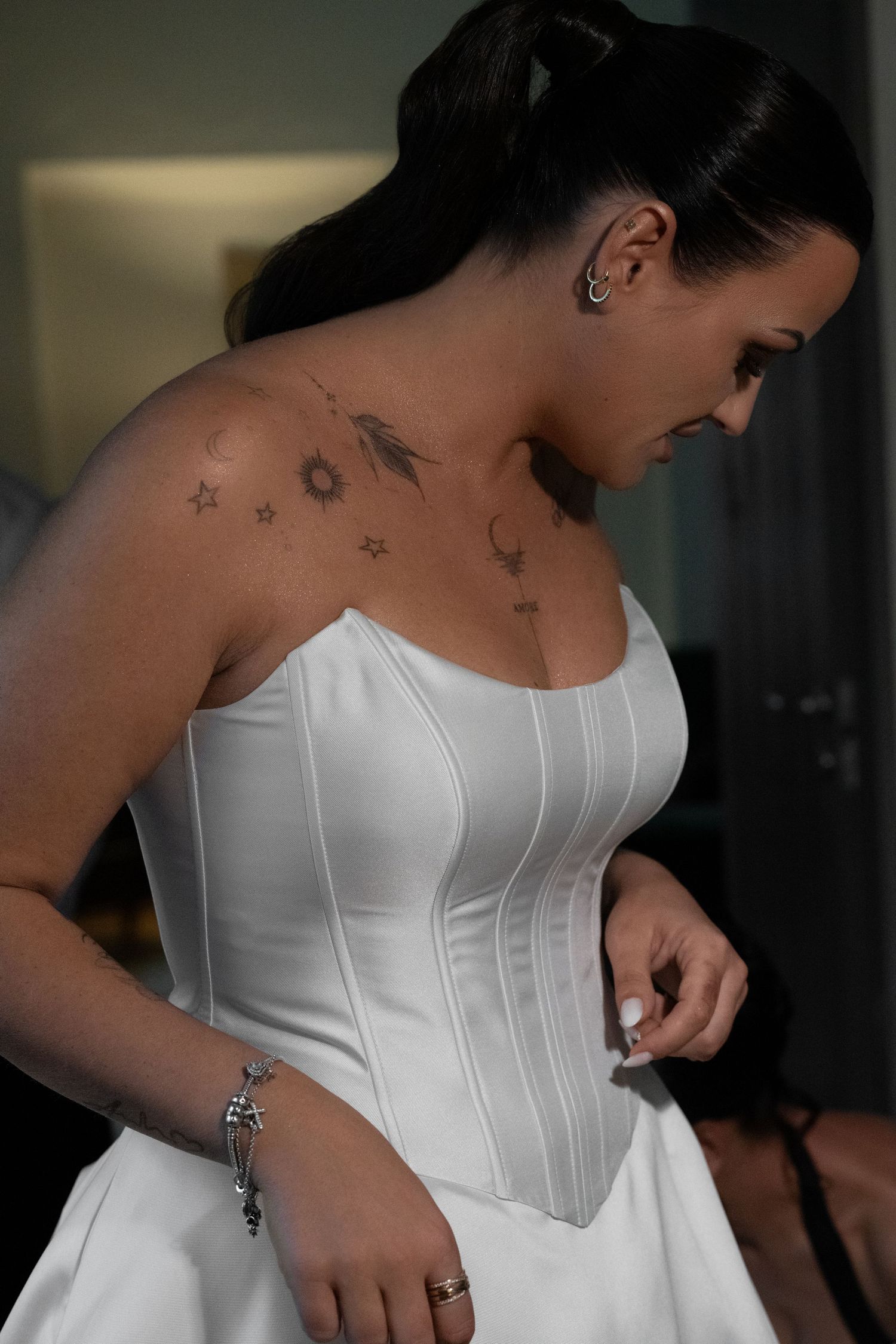 Bride in a white corset-style wedding dress looks down while getting ready, showing star and sun tattoos on her shoulder.
