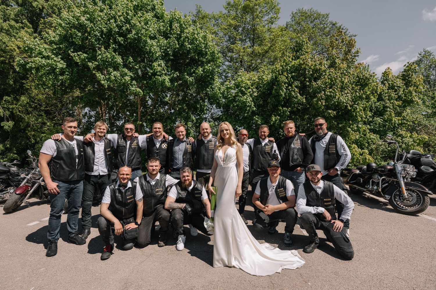 Bride in a white wedding gown poses with a group of men in leather vests beside parked motorcycles under green trees.