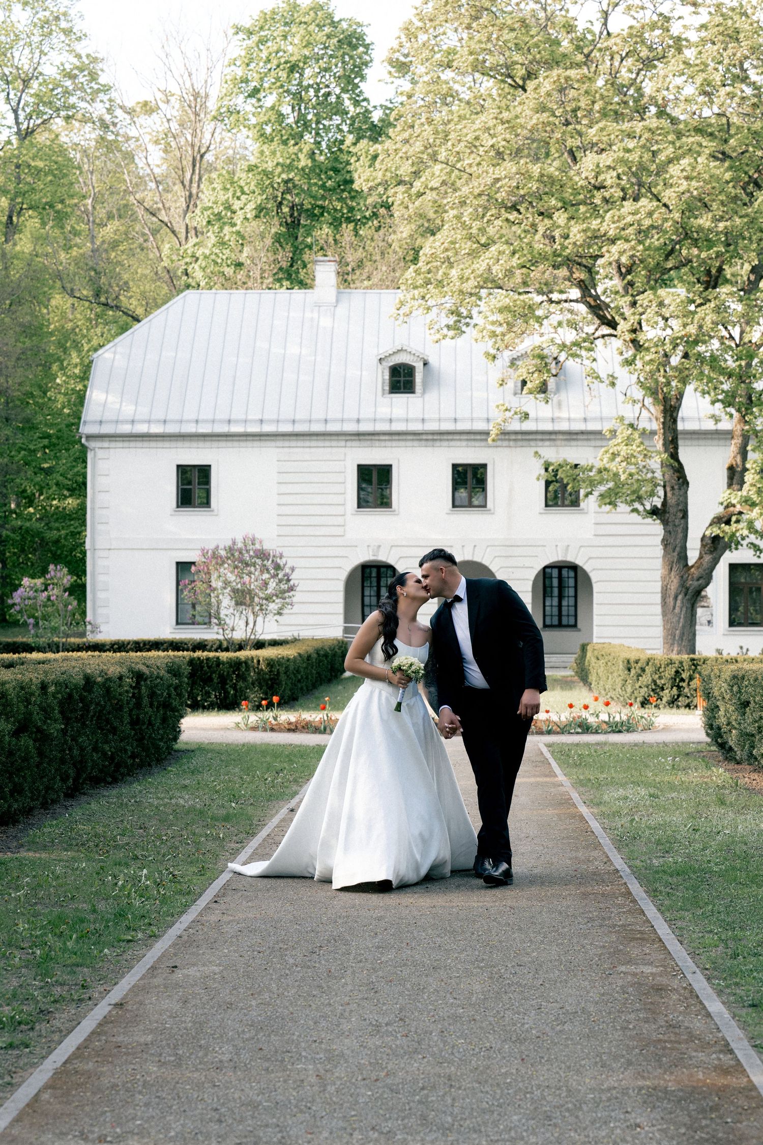 Bride and groom sharing a kiss on a garden pathway in front of a white estate house surrounded by lush green trees.