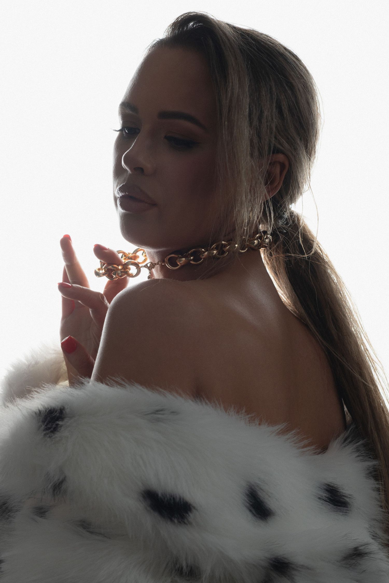 Woman in white fur coat with gold chain choker necklace poses elegantly against bright white background.