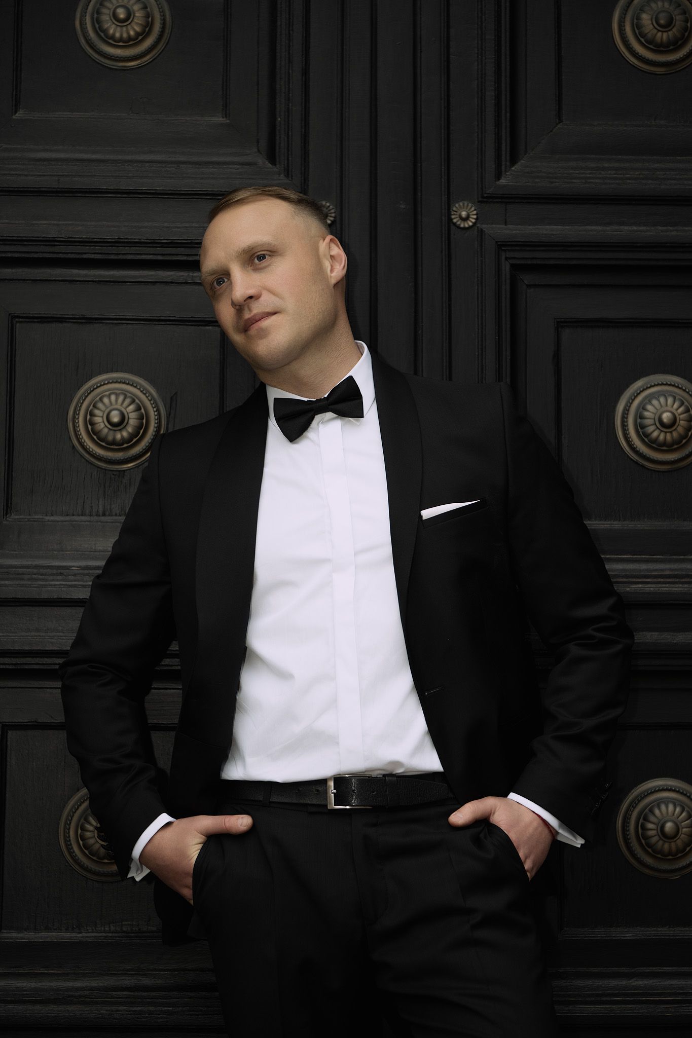 A person wearing a classic black tuxedo and bow tie poses against a dark wooden door with ornate brass details.