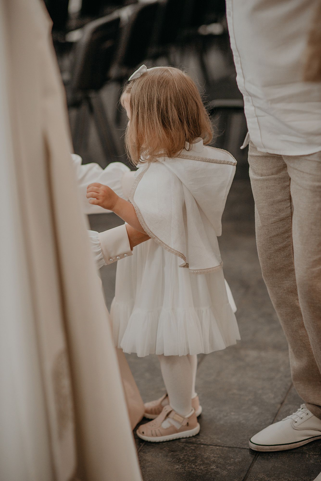 Small child in a white dress and shoes walks between adults wearing white and beige at an elegant event.