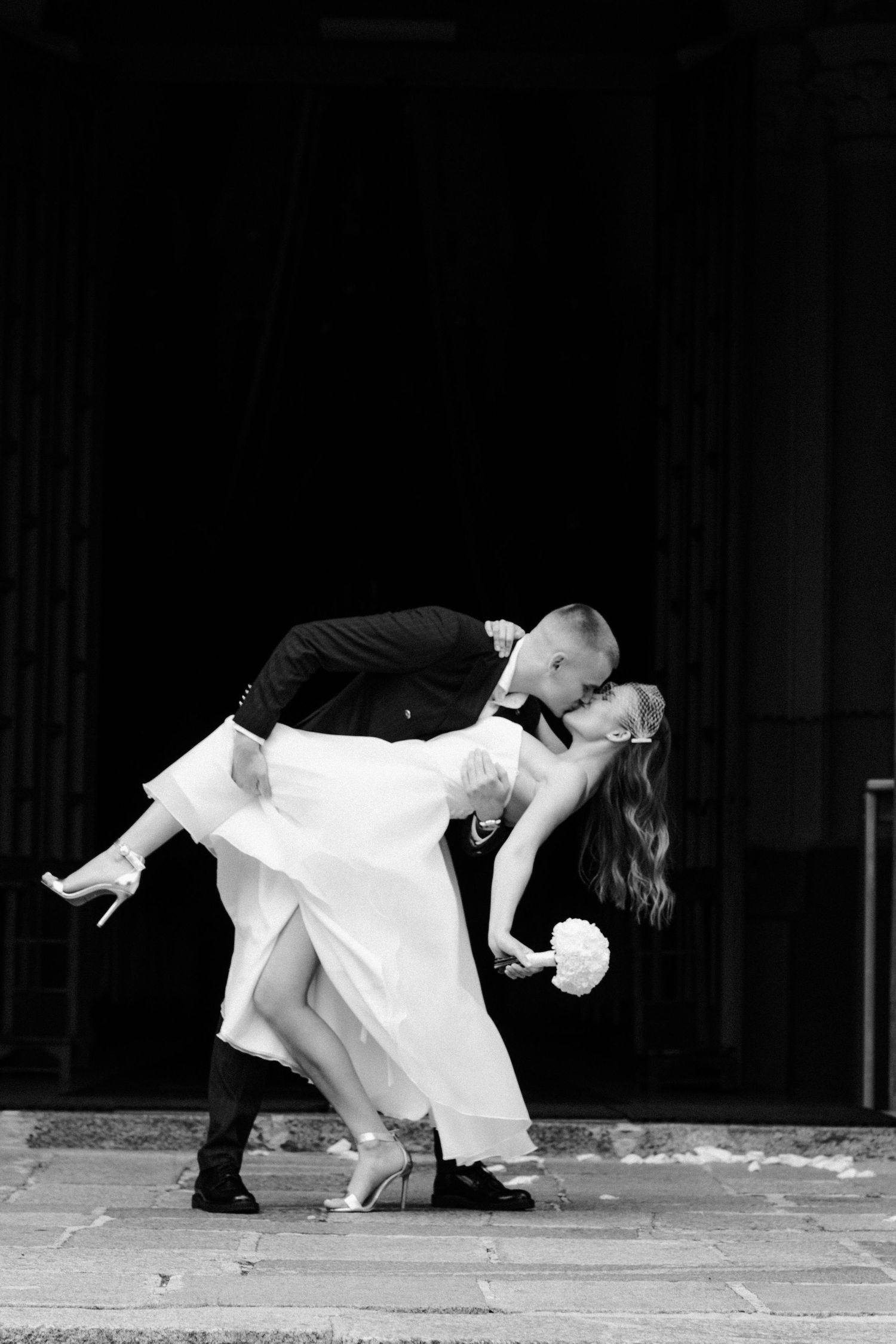 Black and white photo of a bride and groom sharing a romantic dip kiss on stone steps, the bride holding a bouquet.