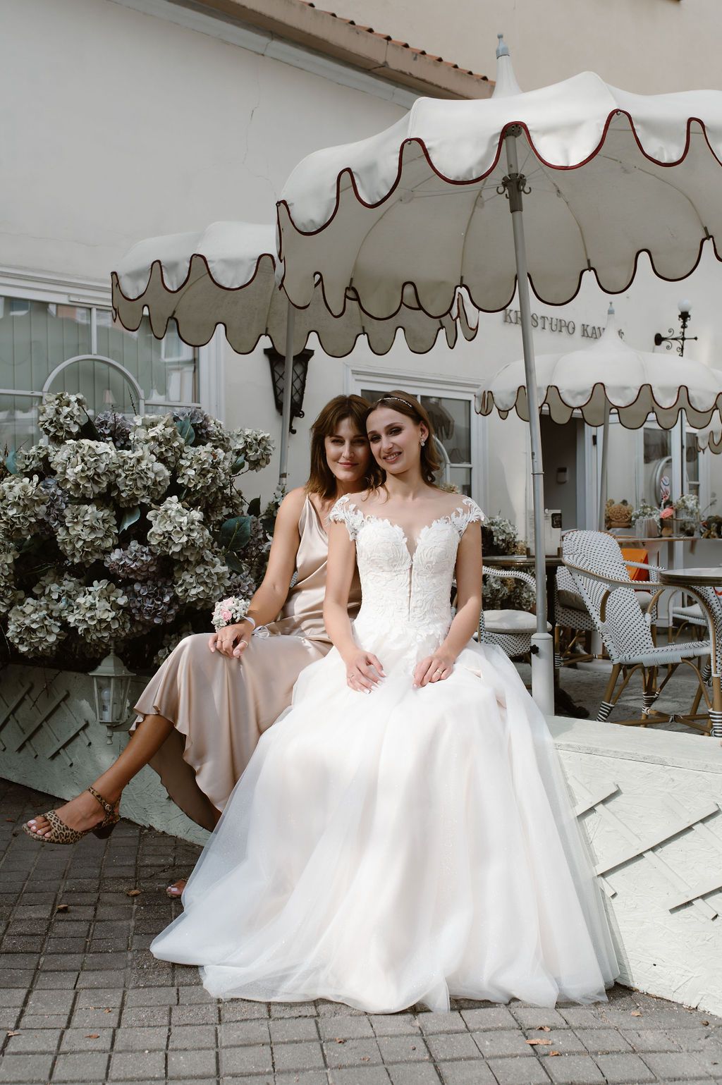 Bride in white ball gown sits with bridesmaid in champagne dress under decorative umbrella at outdoor cafe.