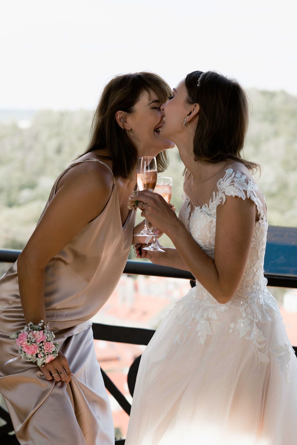 Two women in wedding attire share a kiss while toasting with champagne glasses on a balcony.