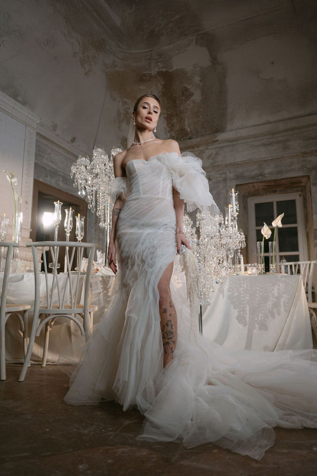 Bride in elegant off-shoulder white tulle mermaid gown with feather sleeves and thigh slit in ornate chandelier venue.