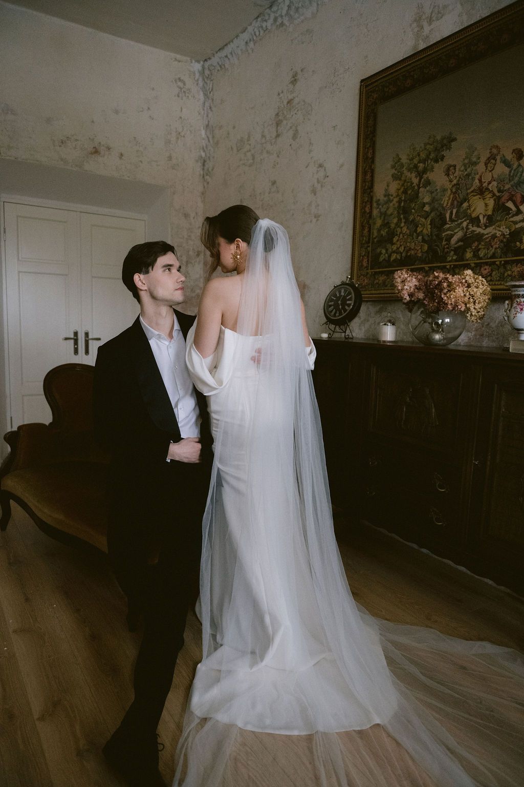 Bride in white gown and veil stands with groom in black suit in elegant vintage room with tapestry.