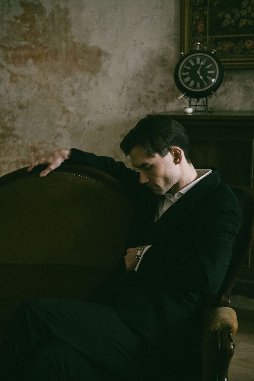Young man in dark suit resting on ornate armchair in moody vintage room with antique clock.