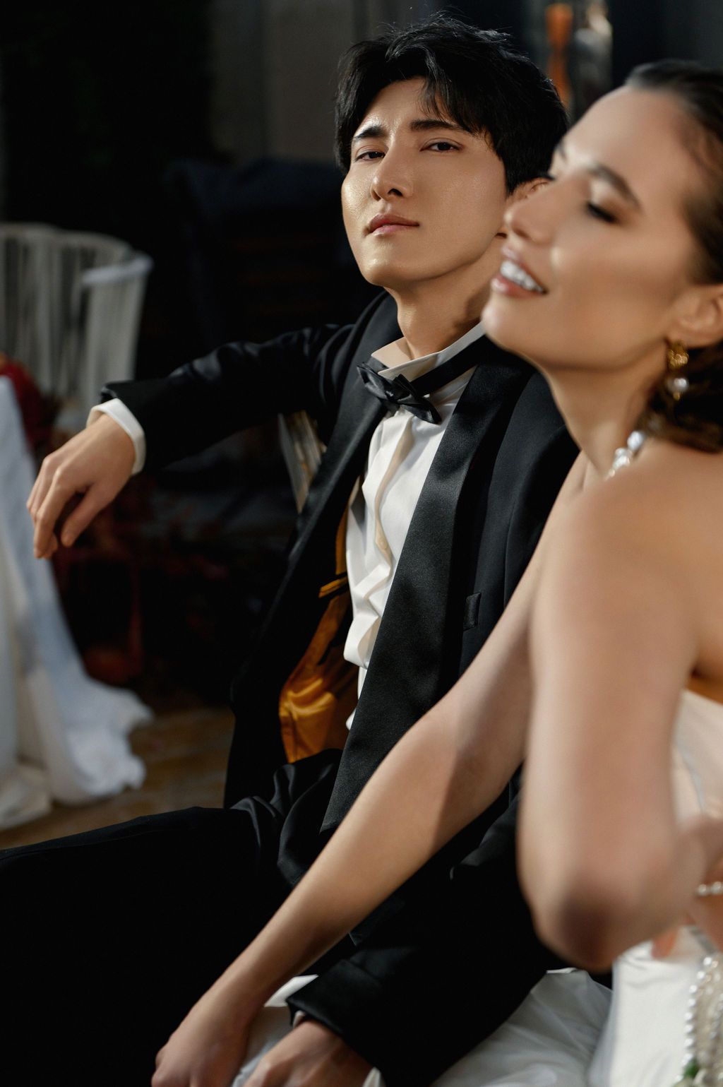 A groom in a black tuxedo with gold sash sits beside a smiling bride in a white dress at a wedding.