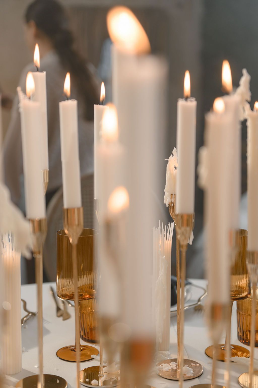 White taper candles in gold candlesticks glowing warmly on a white table at an elegant event.