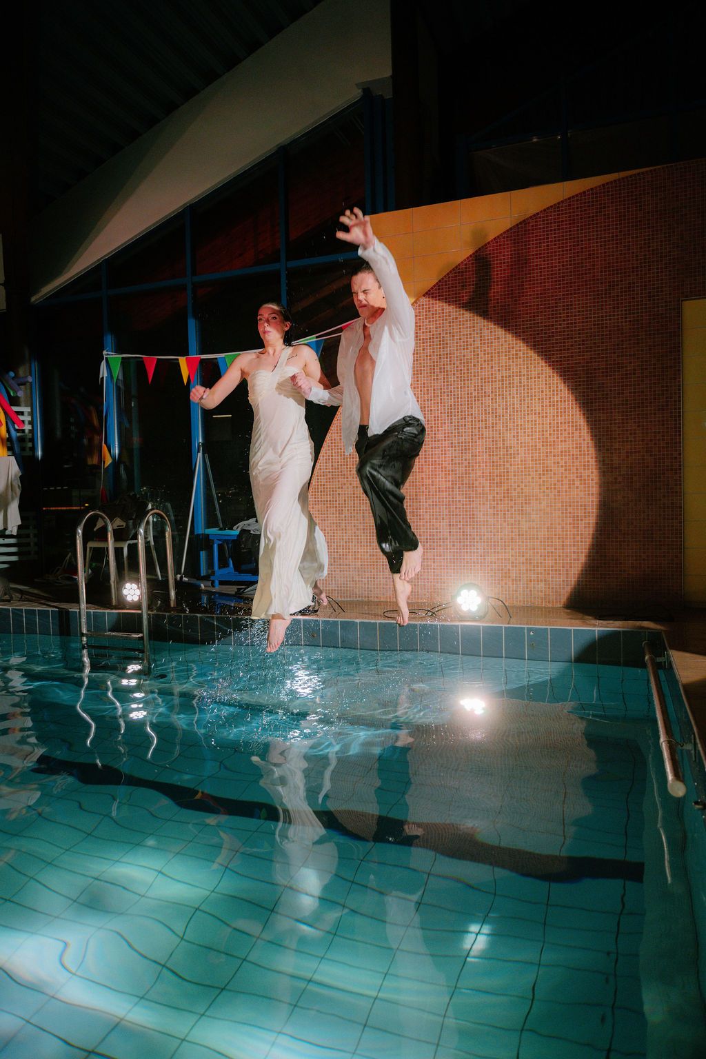 Bride and groom jumping into indoor pool in wedding attire, splashing water in a festive venue.