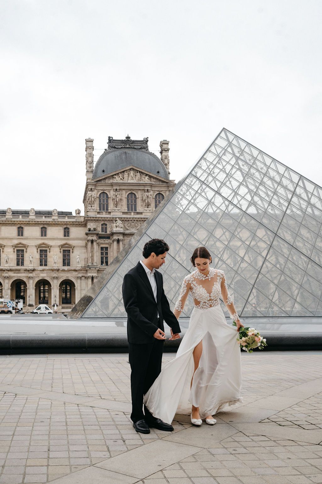 Bride and groom holding hands in front of the Louvre Pyramid in Paris, bride wearing a lace long-sleeve wedding gown.