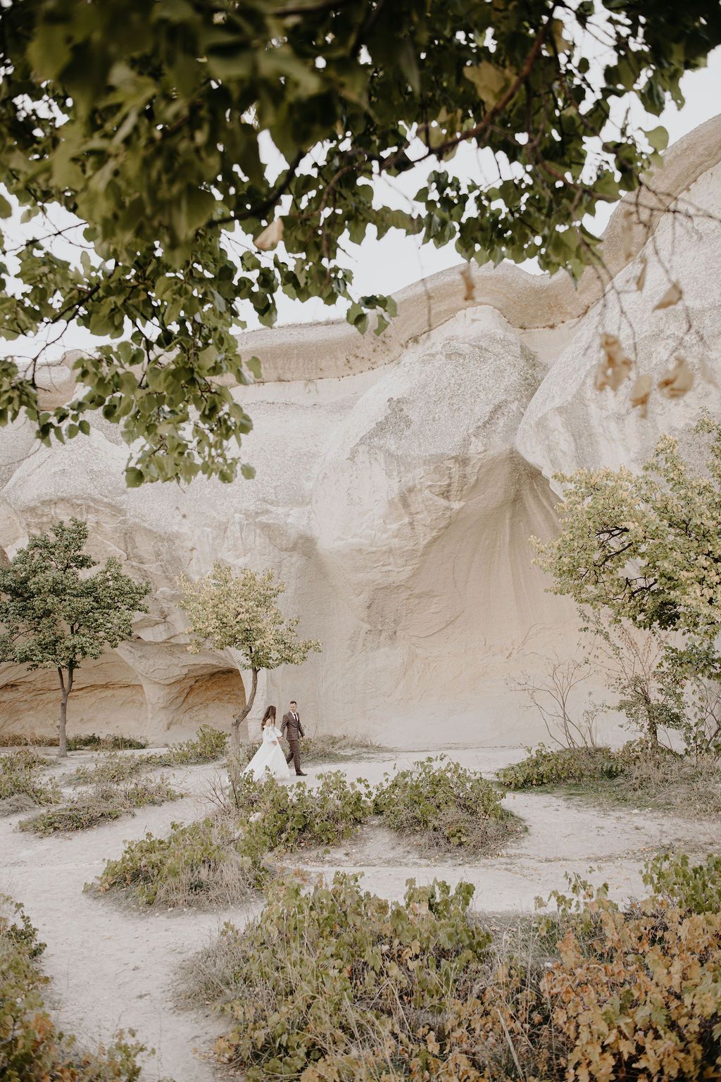 Couple in wedding attire standing among white rock formations and green shrubs in Cappadocia, Turkey.