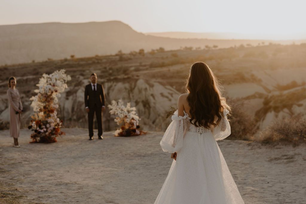 Bride in white gown walks toward groom at desert wedding ceremony during golden sunset in Cappadocia.