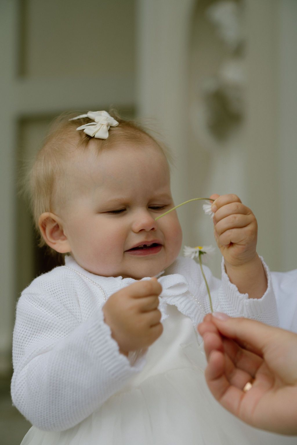 Adorable baby girl in white dress holding a delicate white flower, with a bow in her hair.