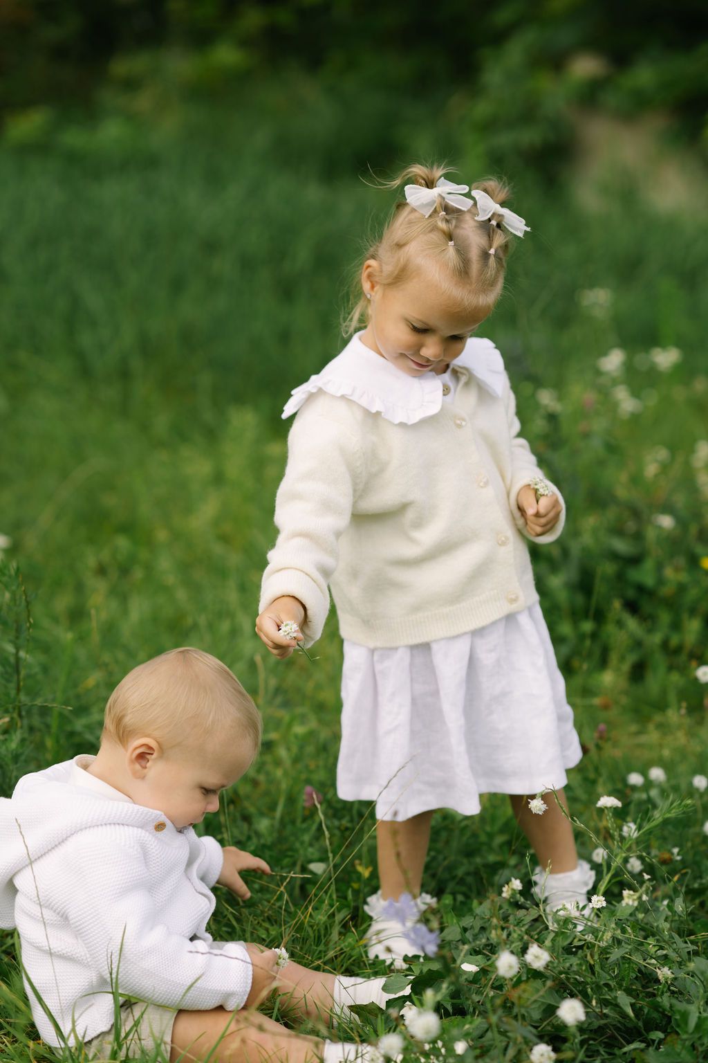 Two young children in white outfits play among wildflowers in a lush green meadow on a sunny day.